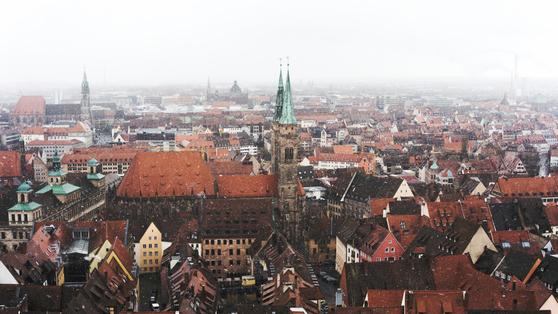 Elevated winter view of Nuremberg&rsquo;s Old Town in Germany, with snow falling across red-tiled rooftops. The twin green spires and large roof of St. Sebald Church dominate the historic cityscape.