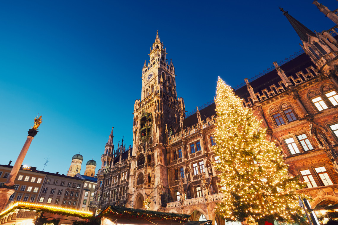 Low-angle evening view of an illuminated Christmas tree outside the New Town Hall at Marienplatz in Munich, Germany. The Mariens&auml;ule and the twin domes of the Frauenkirche are visible beyond the festive square.