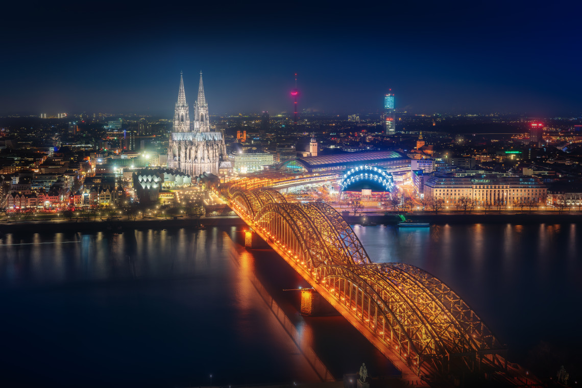 Nighttime aerial view of Cologne Cathedral and the illuminated Hohenzollern Bridge crossing the Rhine River in Cologne, Germany. City lights reflect across the water while the cathedral&rsquo;s twin Gothic towers rise beside the central railway station.