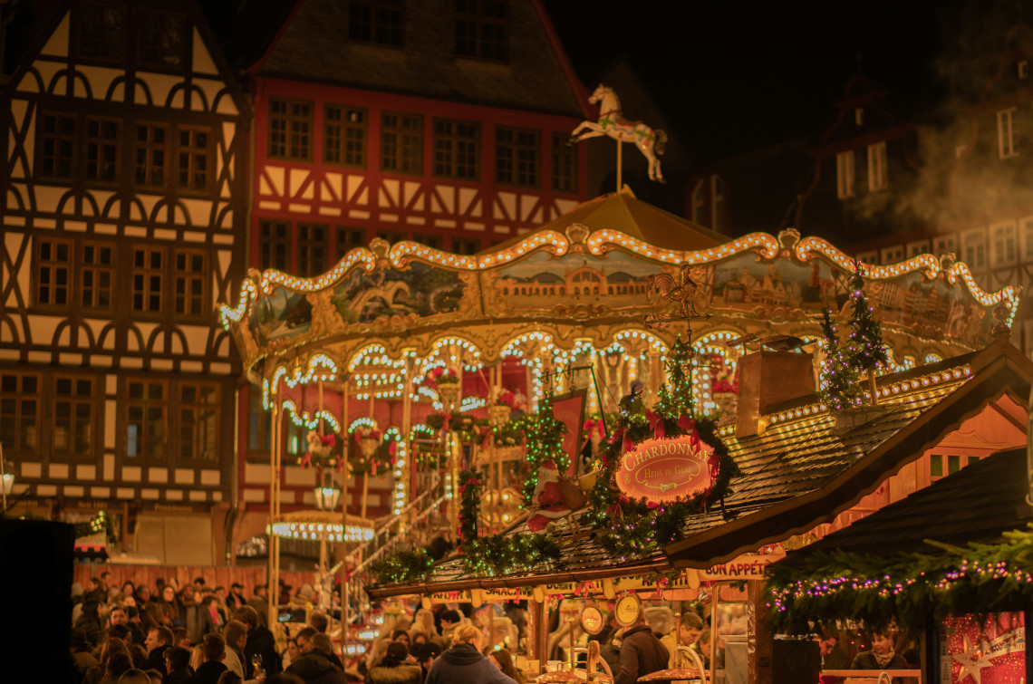 An illuminated traditional carousel rises above the Frankfurt Christmas Market at R&ouml;merberg in Frankfurt, Germany. Festive wooden stalls and visitors fill the square, surrounded by the distinctive half-timbered houses of the historic city centre.