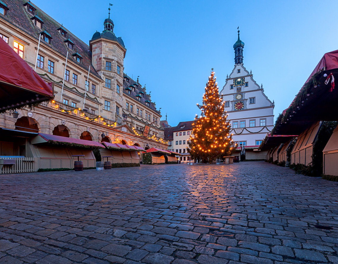 A decorated Christmas tree stands among wooden market stalls in the cobbled Marktplatz of Rothenburg ob der Tauber, Germany. The historic town hall and surrounding medieval buildings frame the Reiterlesmarkt at dusk.