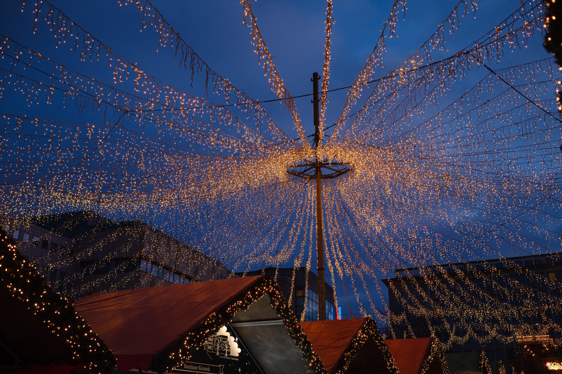 Thousands of warm lights form a canopy above wooden stalls at Dortmund Christmas Market in Dortmund, Germany. The illuminated decorations stretch across the market against the blue evening sky.