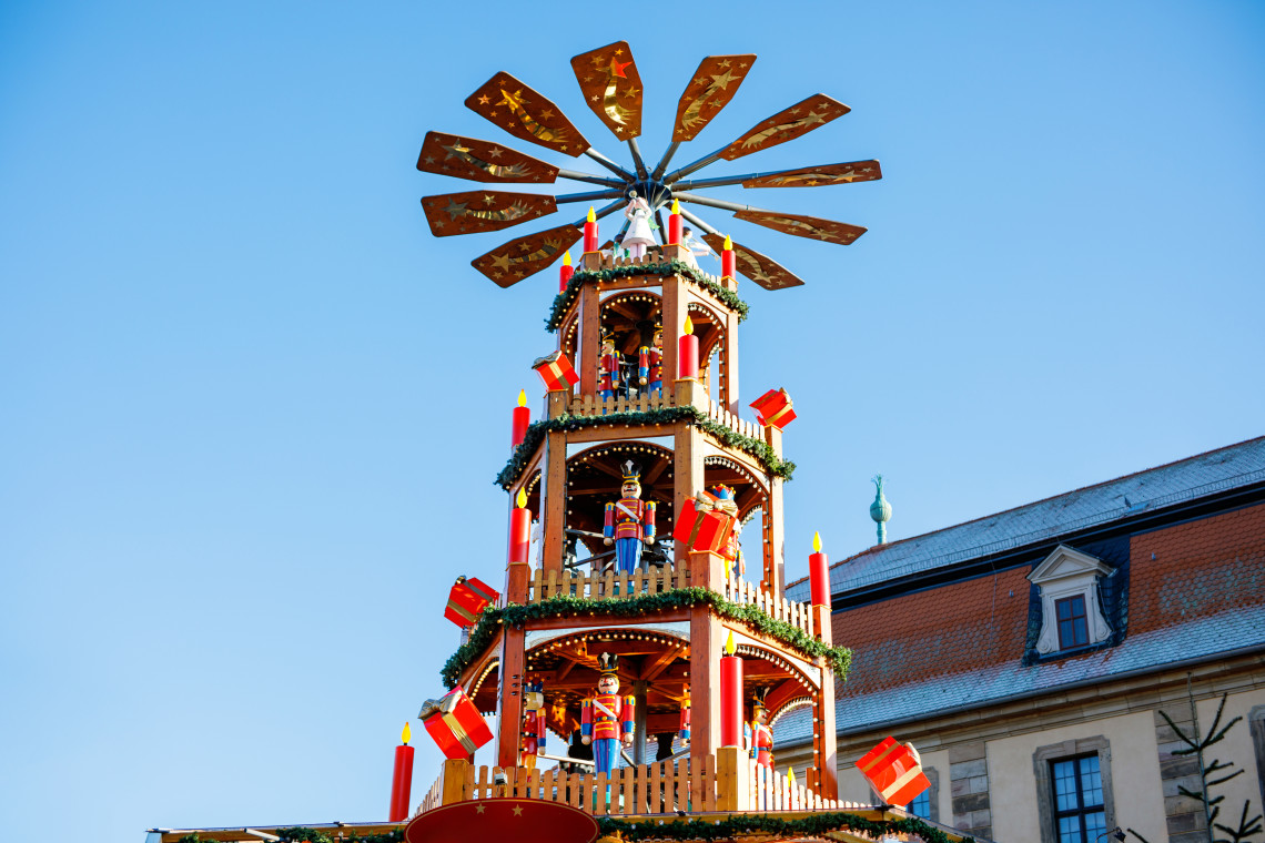 A multi-level wooden Christmas pyramid decorated with candles, garlands and carved figures stands at a Christmas market in Germany. Large rotating blades rise above the traditional structure against a clear blue sky.