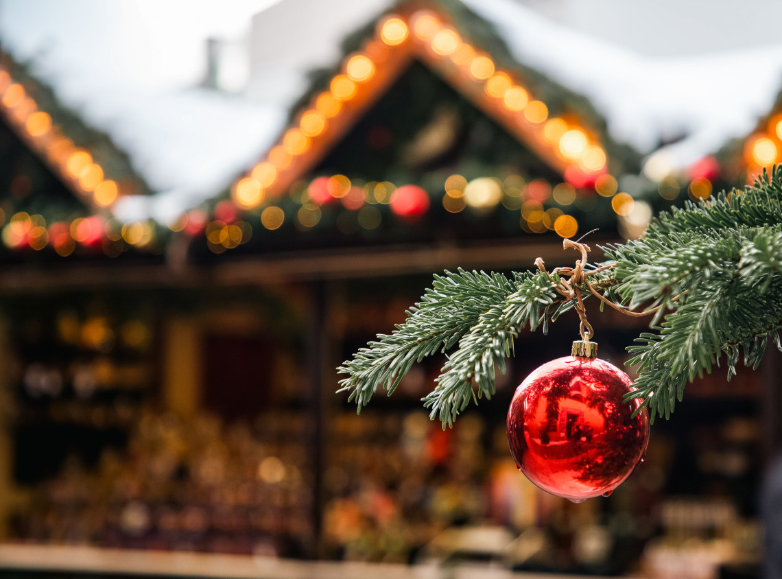 A red Christmas ornament hangs from an evergreen branch at a Christmas market in Germany. Illuminated wooden stalls and festive garlands appear softly blurred in the background.