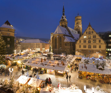 Elevated winter view of the Stuttgart Christmas Market at Schillerplatz in Stuttgart, Germany, with snow-covered stalls and illuminated decorations. The historic Stiftskirche and surrounding Old Town buildings rise behind the festive square.