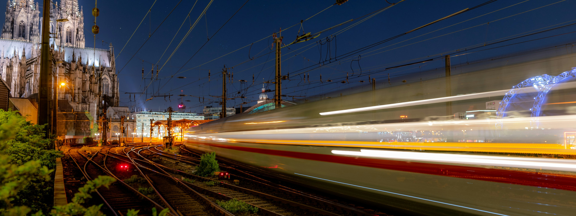 A high-speed train passes Cologne Central Station at night, creating light trails beside Cologne Cathedral in Cologne, Germany. Railway tracks and overhead power lines fill the foreground.