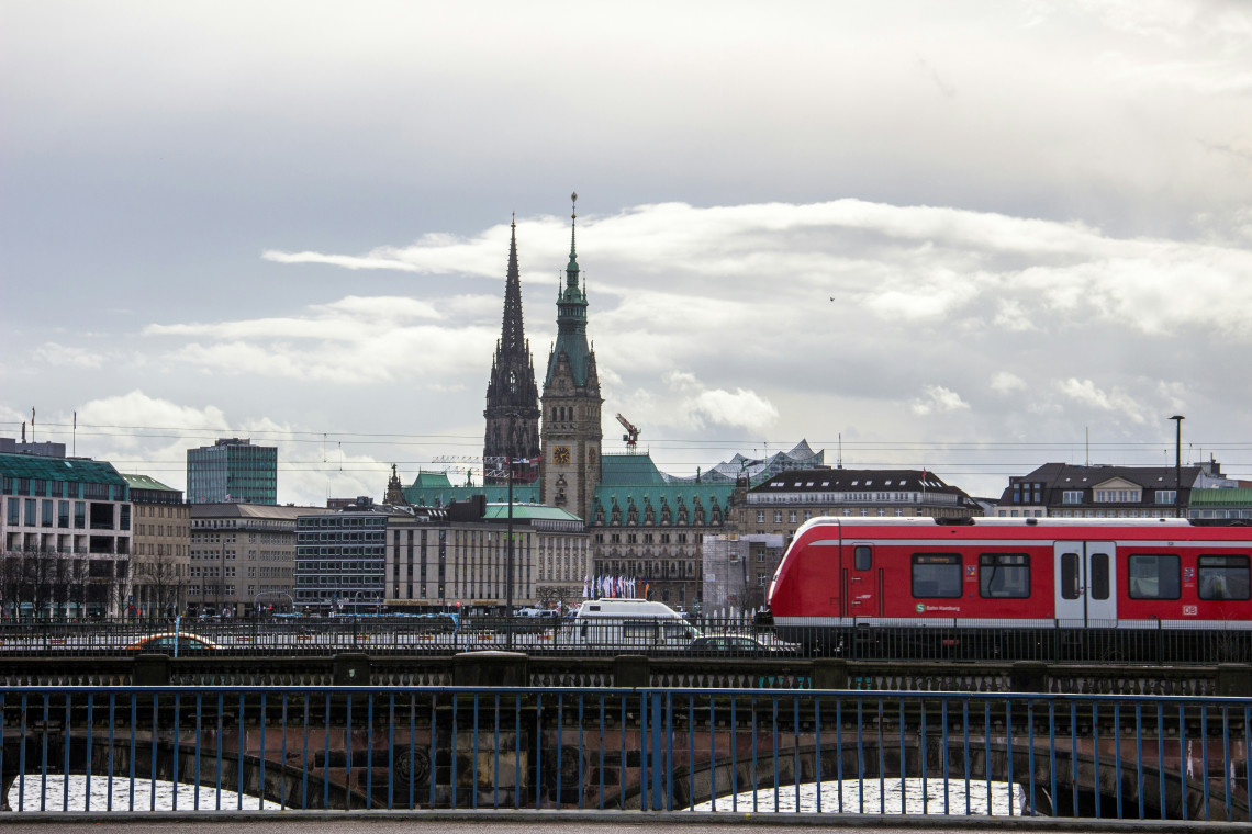 A red S-Bahn train crosses central Hamburg, Germany, with historic church towers and city buildings rising beneath a cloudy sky.