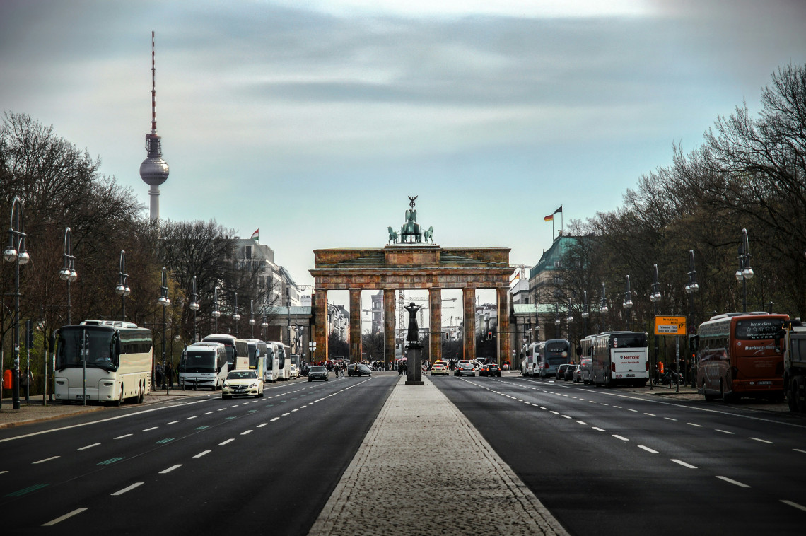 The Brandenburg Gate stands at the end of Unter den Linden in Berlin, Germany, with the Berlin TV Tower visible to the left. Buses and vehicles line the broad avenue leading towards the landmark.