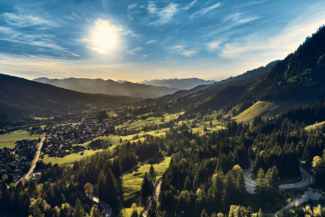 Sunlight spreads across a green valley in the Bavarian Alps, Germany, surrounded by forested mountain slopes and distant peaks. A winding road curves through the hillside above a small village.