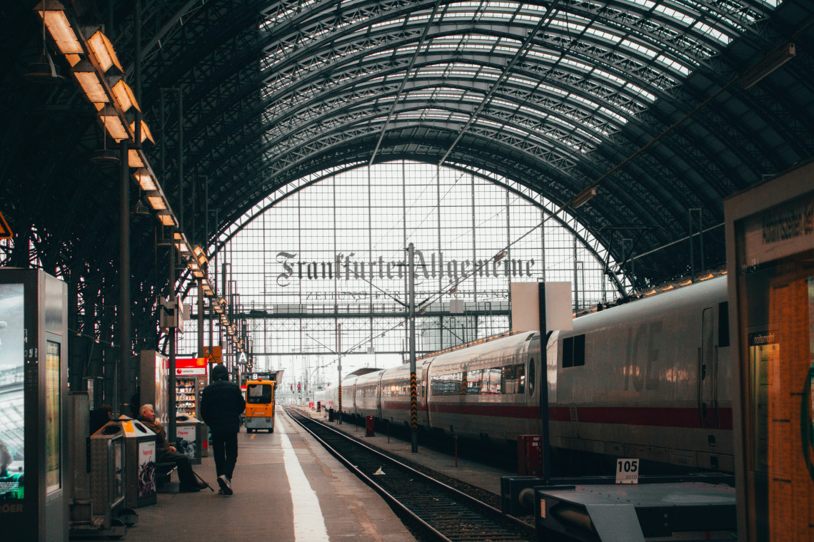 An ICE high-speed train waits beside a platform inside Frankfurt Central Station in Frankfurt, Germany. The station&rsquo;s arched glass-and-steel roof frames the railway tracks and passenger area.