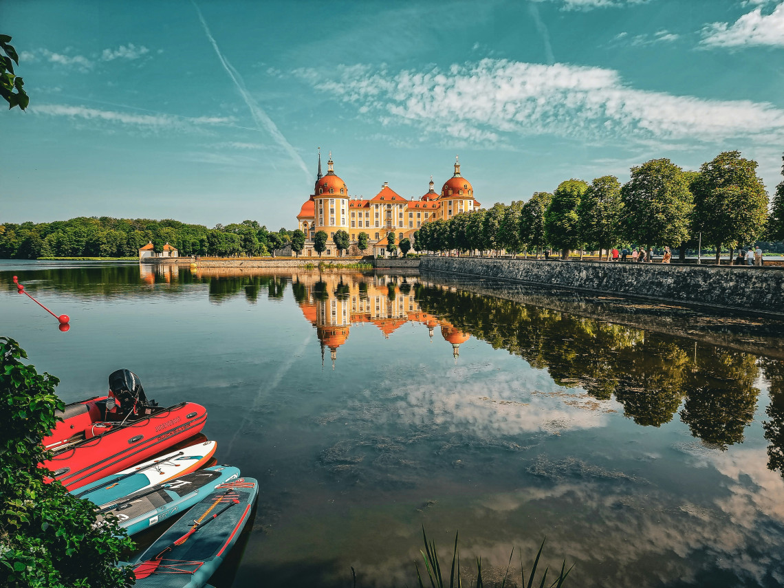 Moritzburg Castle is reflected in the calm lake surrounding the Baroque palace in Moritzburg, Germany. Paddleboards and a small red boat rest near the tree-lined shoreline in the foreground.  Image 14