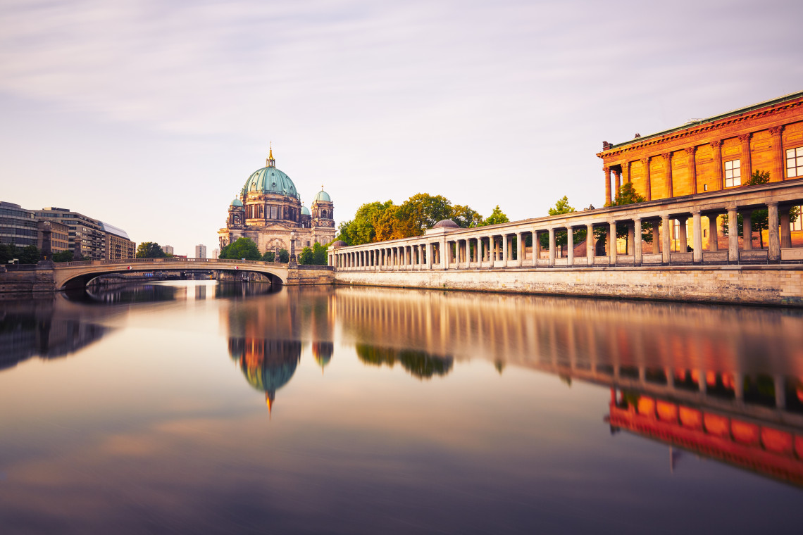 Berlin Cathedral rises beside the Spree River on Museum Island in Berlin, Germany. The cathedral dome, riverside colonnade and surrounding museum buildings are reflected in the still water.