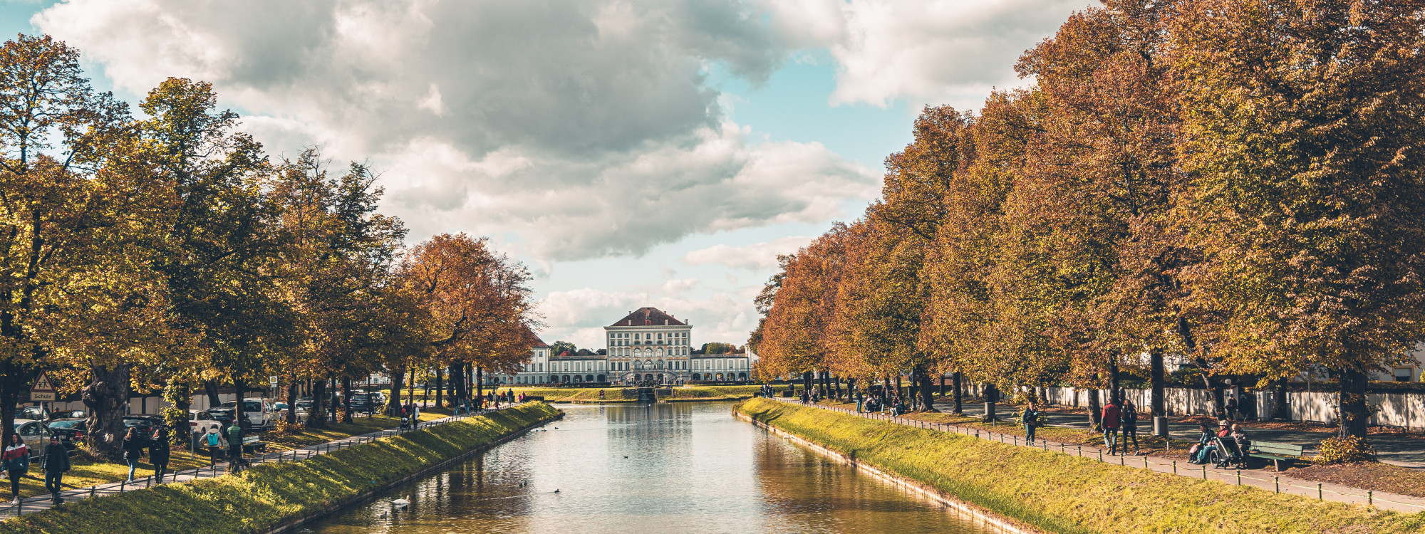 The canal leads toward Nymphenburg Palace in Munich, Germany, bordered by rows of trees with autumn foliage. Pedestrians and cyclists follow the paths beside the water.