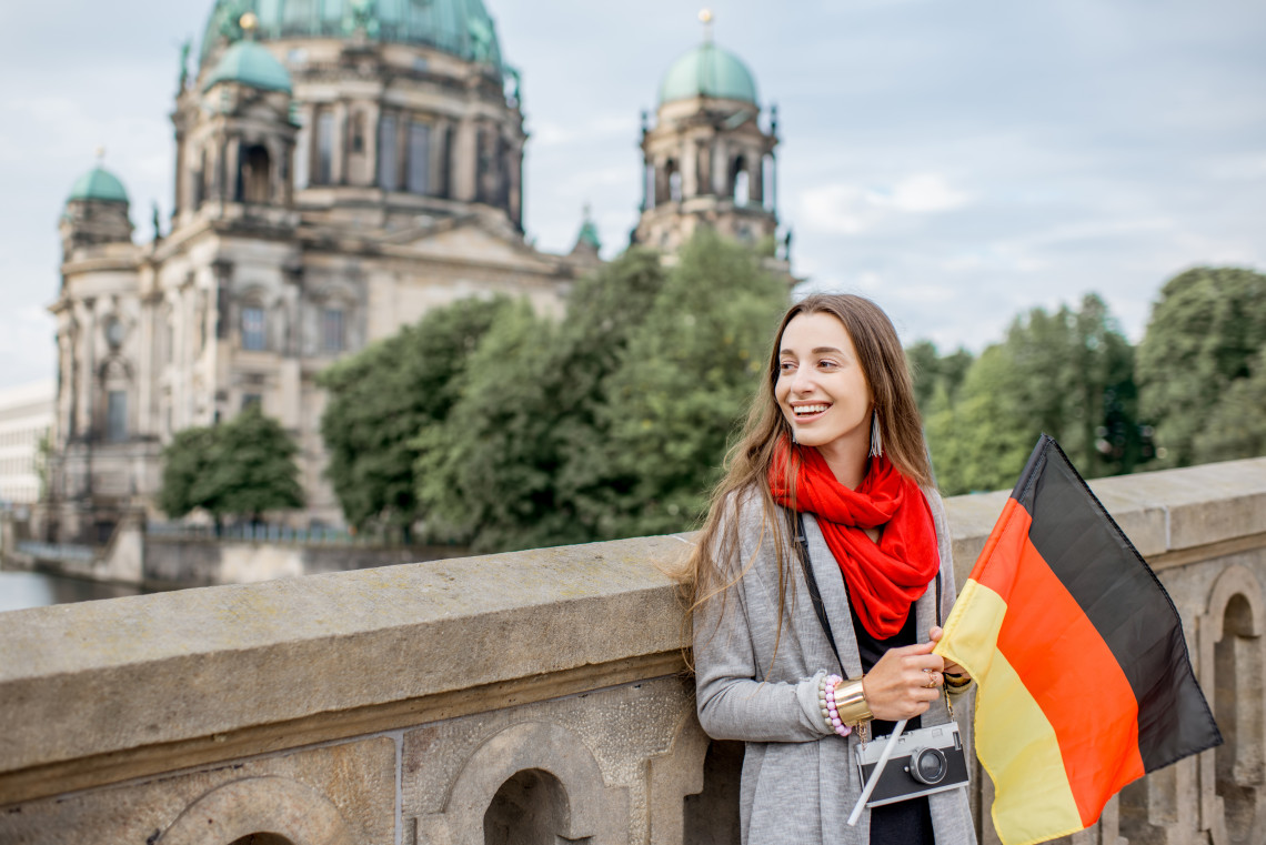 A traveler holds the German flag near Berlin Cathedral in Berlin, Germany. The cathedral&rsquo;s stone fa&ccedil;ade and green copper domes appear behind her.
