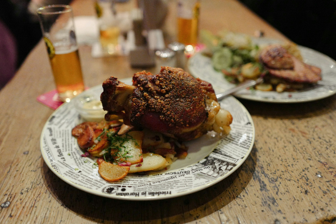 A roasted Bavarian pork knuckle with a seasoned crust is served over sliced potatoes at a restaurant in Munich, Germany. A glass of beer and a second meal sit on the wooden table.