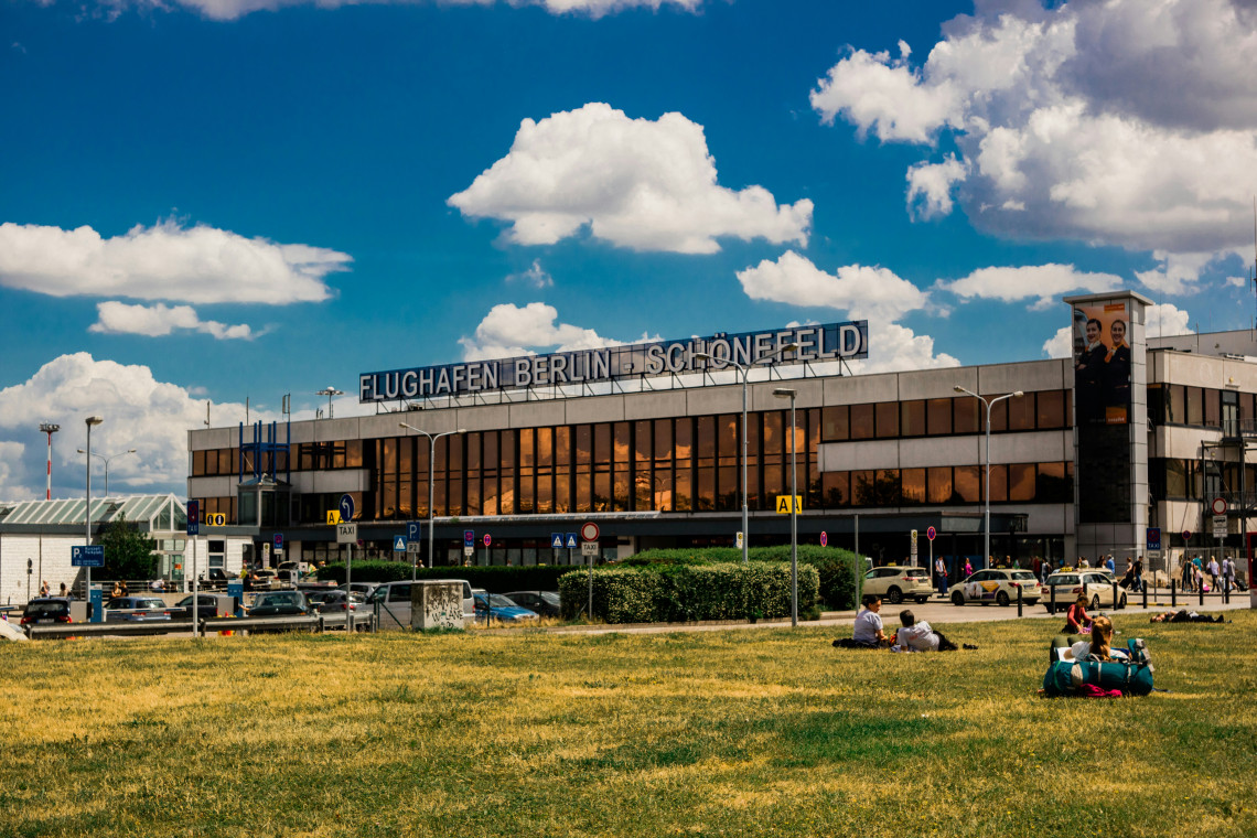 The former Berlin Sch&ouml;nefeld Airport terminal in Berlin, Germany, displays its original rooftop signage above the entrance. Cars, travelers and a grassy public area fill the foreground.