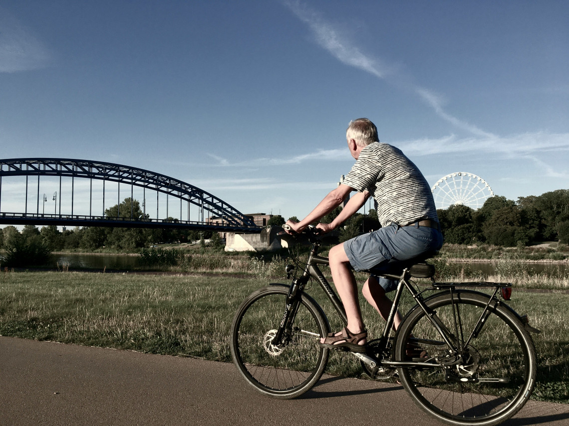 A man cycles along a paved riverside path in Berlin, Germany, with a steel arch bridge and Ferris wheel visible in the background.