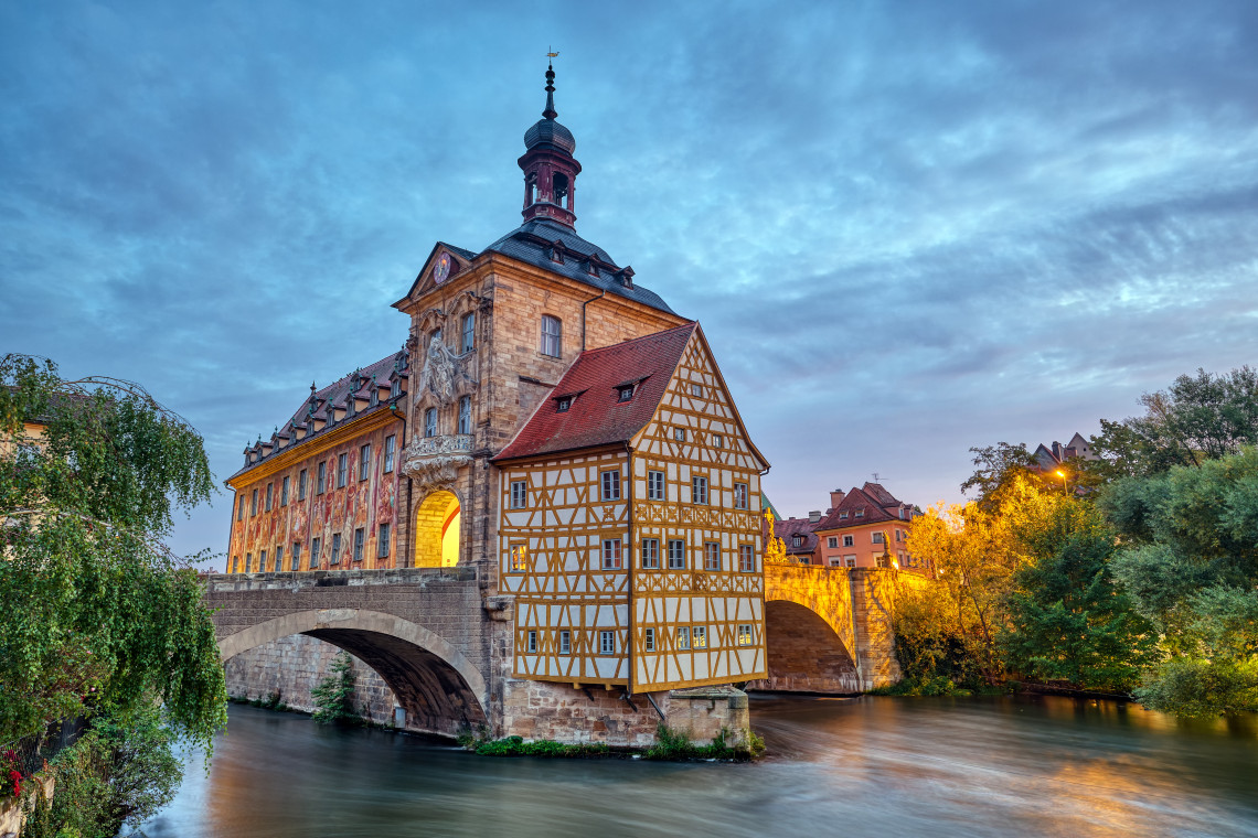Bamberg&rsquo;s Old Town Hall stands on a stone bridge above the Regnitz River in Bamberg, Germany. Its Baroque fa&ccedil;ade and half-timbered section are illuminated beneath a cloudy evening sky.