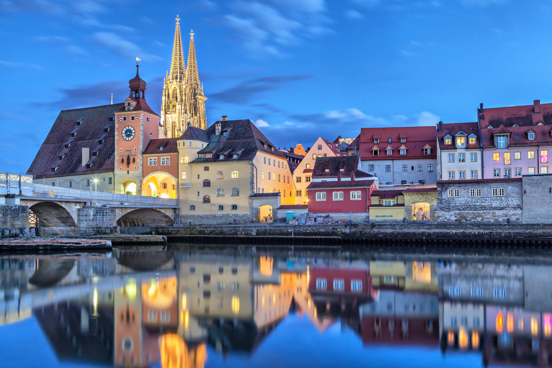 Regensburg Cathedral and the historic bridge tower rise above the Danube River in Regensburg, Germany. Illuminated Old Town buildings are reflected in the water at dusk.
