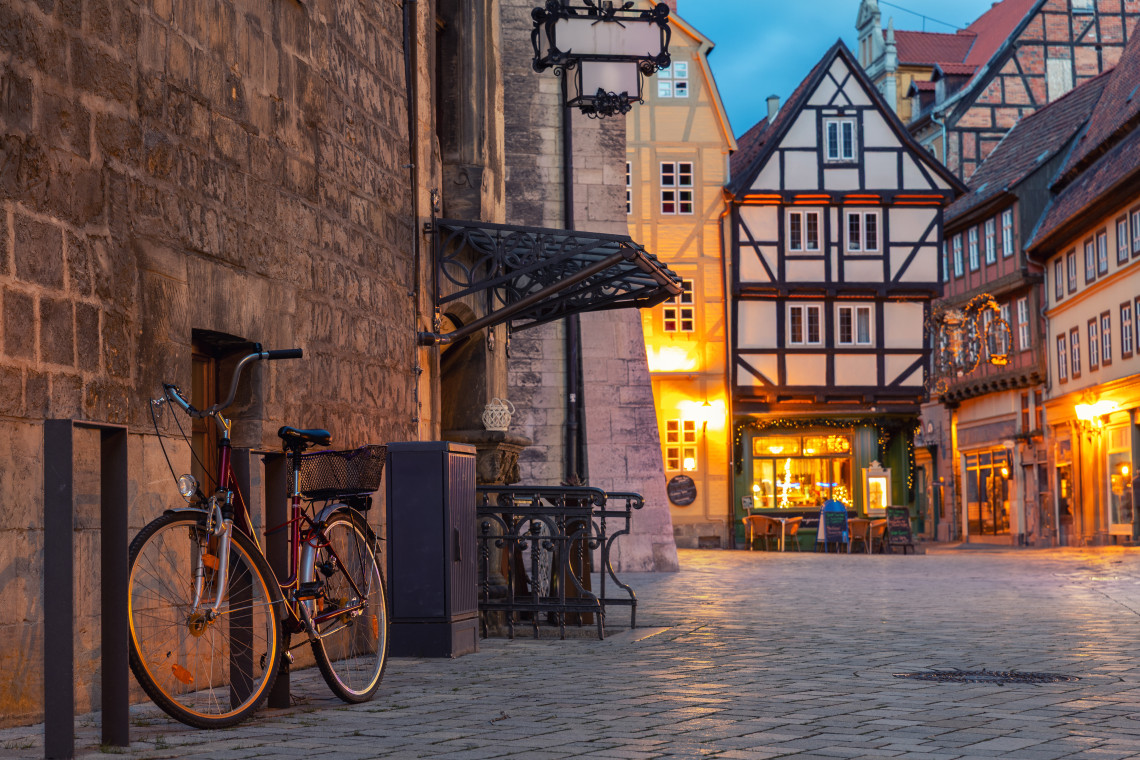 A bicycle rests beside a cobblestone street in Quedlinburg Old Town, Germany. Half-timbered houses, historic stone walls and illuminated shopfronts frame the quiet evening scene.