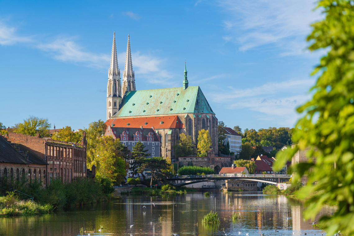 The twin spires and green roof of St Peter and Paul Church rise above the Neisse River in G&ouml;rlitz, Germany. Trees, riverside buildings and a pedestrian bridge surround the Gothic landmark.