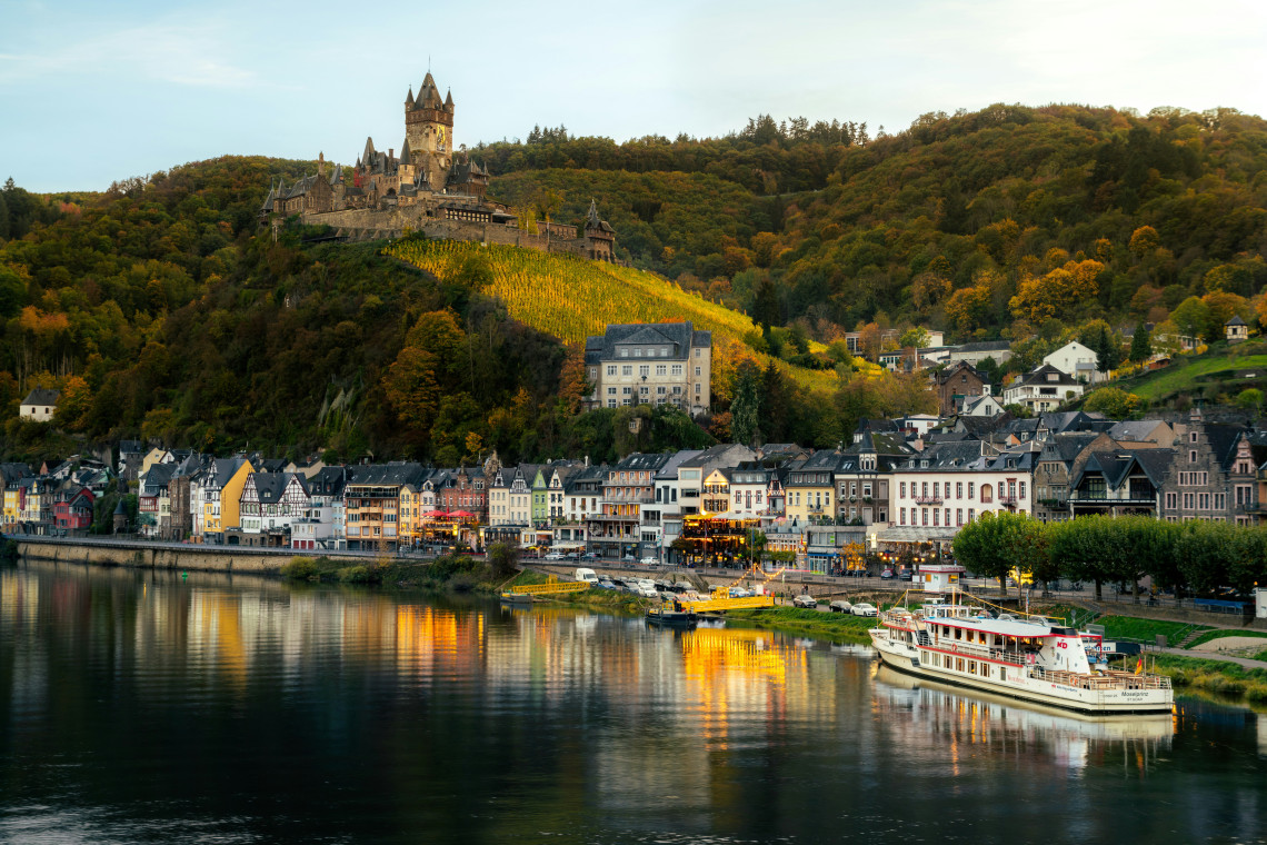 Reichsburg Cochem stands above vineyard-covered hills and the riverside town of Cochem, Germany. Colourful buildings and a passenger boat are reflected in the Moselle River below.