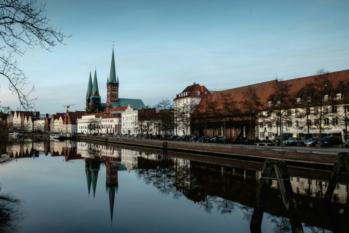 The twin spires of L&uuml;beck Cathedral rise above the historic waterfront in L&uuml;beck, Germany. Old Town buildings and church towers are reflected in the calm waters of the Trave River.