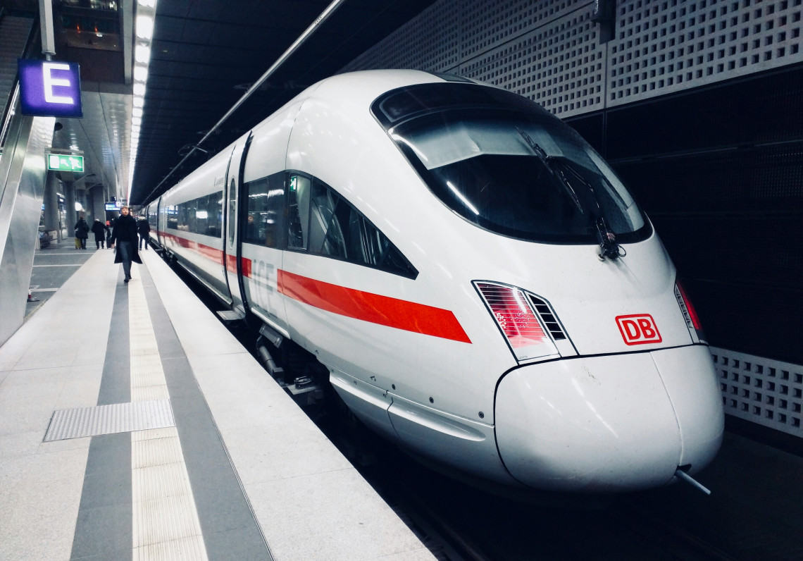 A white Deutsche Bahn ICE high-speed train waits beside a station platform in Berlin, Germany. Its streamlined front, red stripe and passenger carriages are visible beneath the covered railway terminal.