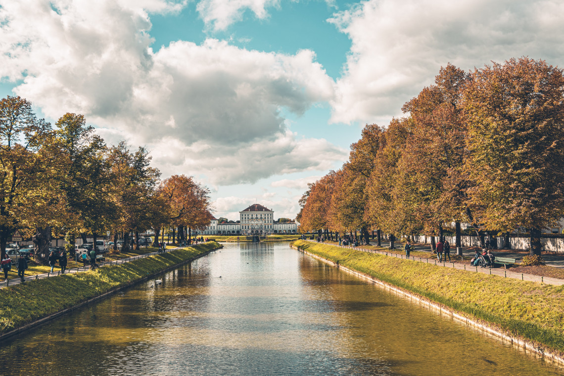 The canal leads towards Nymphenburg Palace in Munich, Germany, bordered by rows of trees displaying autumn foliage. Visitors walk and cycle along the paths beside the water.