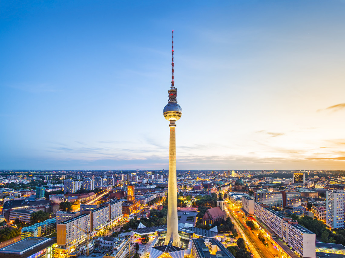 The illuminated Berlin TV Tower stands above the city centre of Berlin, Germany, surrounded by urban buildings and busy streets at dusk.