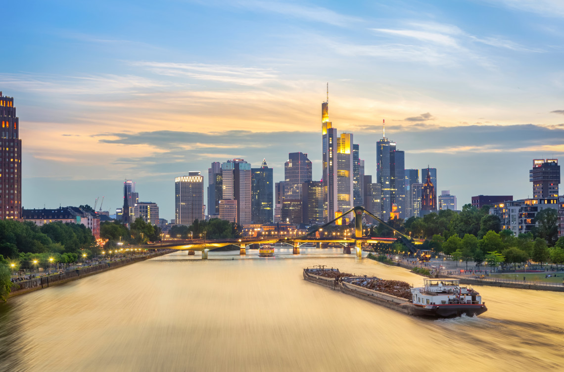 The towers of Frankfurt&rsquo;s financial district rise beyond the Main River in Frankfurt, Germany, as boats travel past illuminated bridges and waterfront paths at sunset.