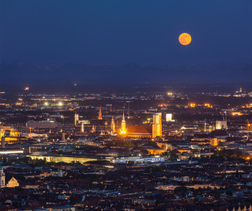 Night view over Munich with illuminated city streets beneath a full moon and the distant Bavarian Alps on the horizon.