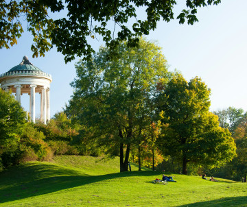 The Monopteros pavilion stands on a grassy hill in the English Garden in Munich, Germany. Mature trees surround visitors relaxing across the sunlit park.