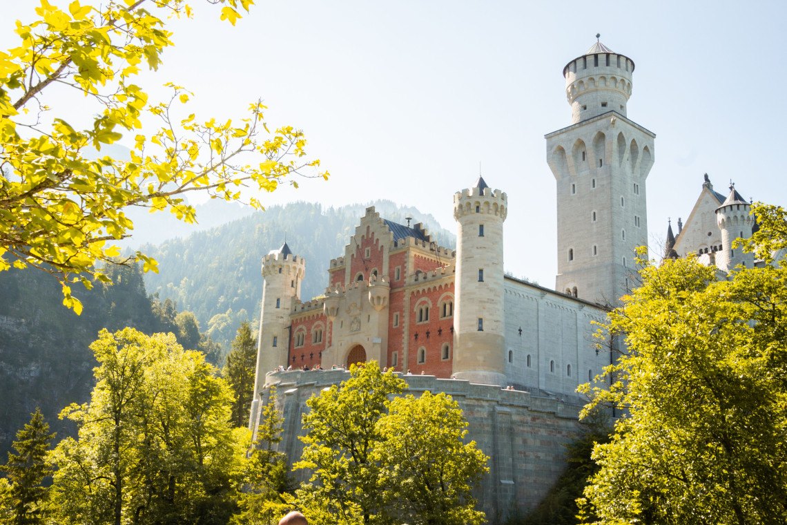 The white towers and red entrance fa&ccedil;ade of Neuschwanstein Castle rise above autumn trees near Schwangau, Germany. Sunlit foliage frames the historic Bavarian landmark.