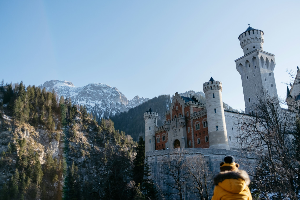 Neuschwanstein Castle stands above a snow-covered forest near Schwangau, Germany. The aerial view reveals its towers, mountain setting and the Bavarian Alps beneath a pink winter sky.