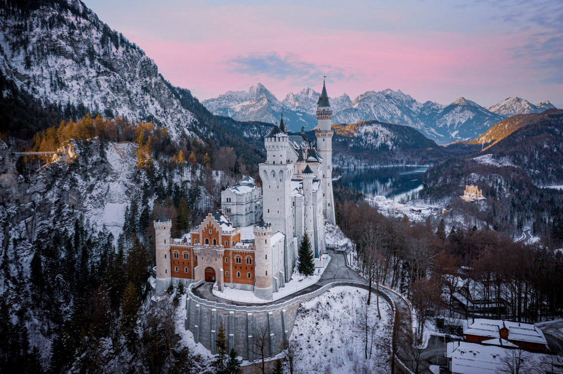 Neuschwanstein Castle appears among forested foothills near Schwangau, Germany, beneath the rocky peaks of the Bavarian Alps. A broad green meadow fills the foreground.