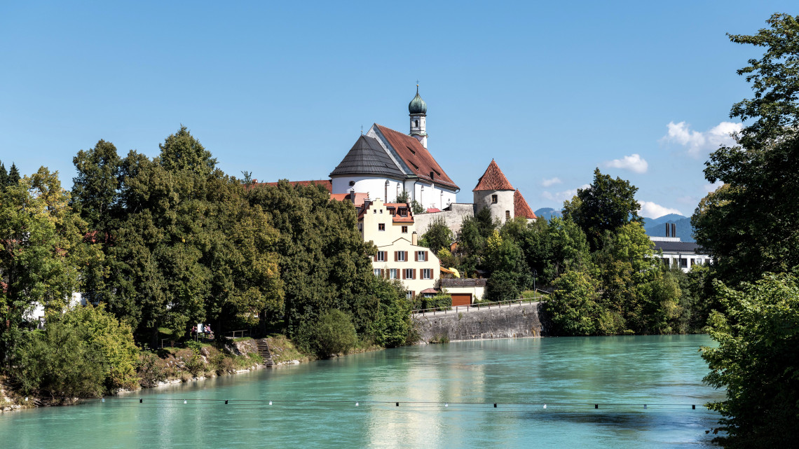 Franziskanerkirche St. Stephan stands above the turquoise Lech River in F&uuml;ssen, Germany. The white church, onion-domed tower and adjoining historic buildings are surrounded by trees.