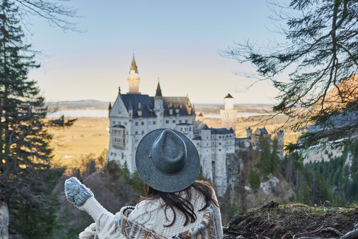 A traveler wearing a wide-brimmed hat sits at a forest viewpoint overlooking Neuschwanstein Castle near Schwangau, Germany. The palace rises above the surrounding Bavarian countryside.
