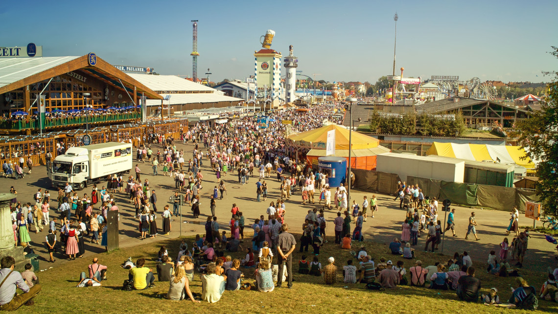 Crowds move between beer tents and fairground attractions at Oktoberfest on Theresienwiese in Munich, Germany. Visitors sit on the hillside overlooking the festival grounds.