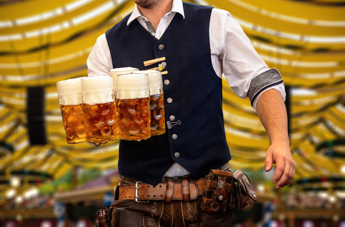 A waiter wearing traditional Bavarian clothing carries several large beer steins inside an Oktoberfest tent in Munich, Germany. The mugs are filled with amber beer and topped with foam.