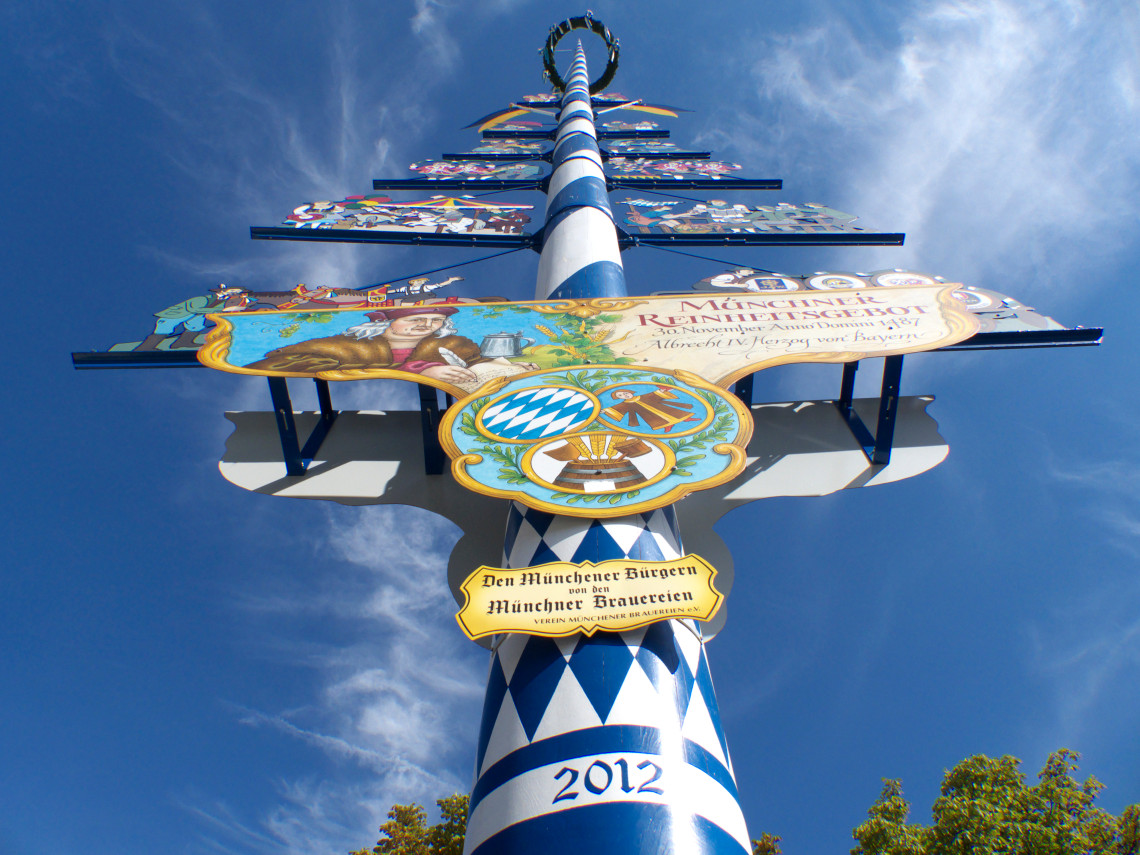 The blue-and-white maypole at Viktualienmarkt in Munich, Germany, is decorated with painted signs representing local trades and Bavarian breweries. The structure rises against a clear blue sky.