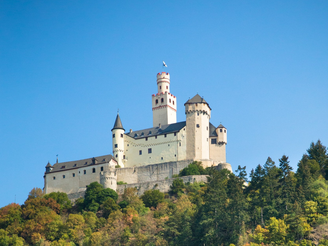 Marksburg Castle rises above a forested hillside in Braubach, Germany. Its pale stone walls, fortified towers and red-and-white keep stand against a clear blue sky.