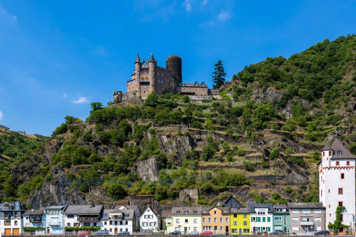 Sch&ouml;nburg Castle stands on a rocky, forested hillside above Oberwesel, Germany. Colourful riverside buildings and a historic tower line the town below.