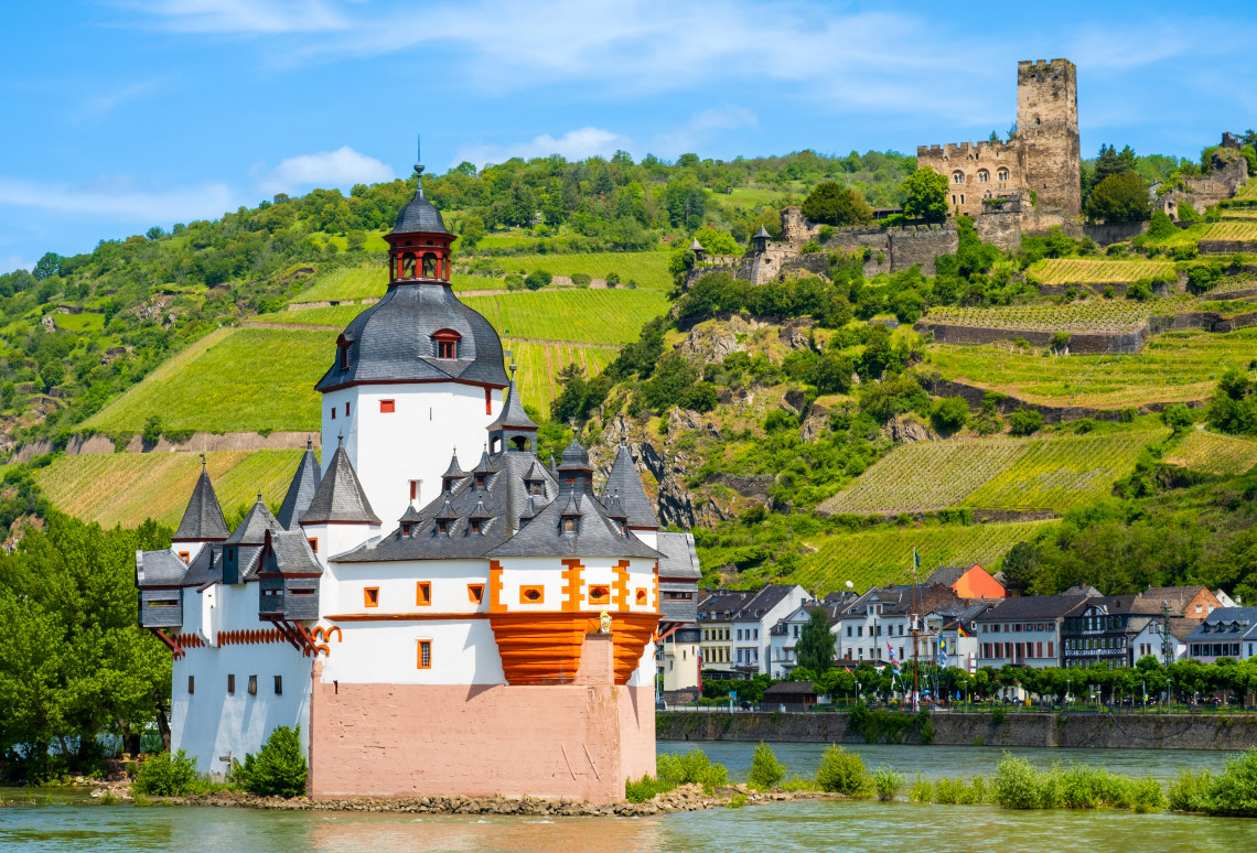Pfalzgrafenstein Castle stands on a small island in the Rhine River at Kaub, Germany. Gutenfels Castle and vineyard-covered hills rise behind the white-and-red customs fortress.