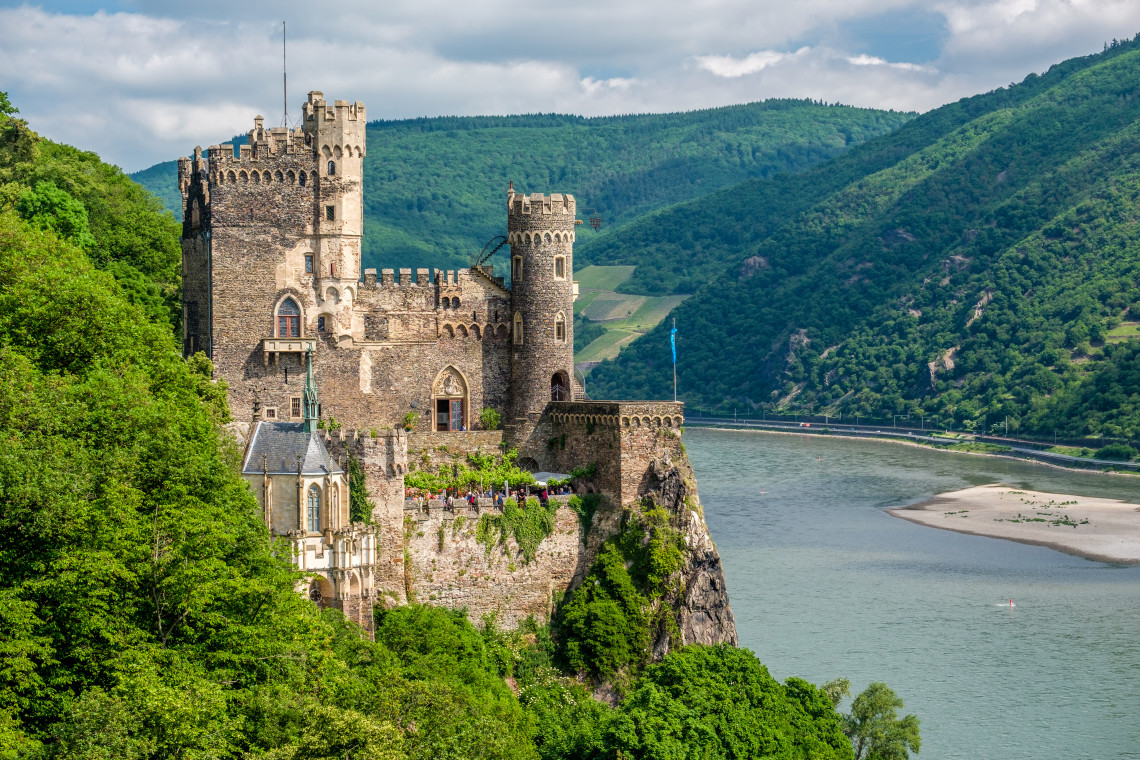 Rheinstein Castle occupies a forested cliff above the Rhine River near Trechtingshausen, Germany. Stone towers, crenellated walls and a small Gothic chapel overlook the river valley.