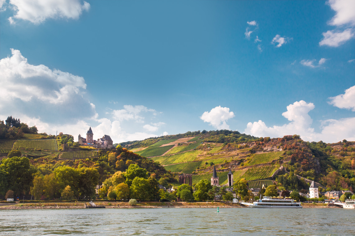 Stahleck Castle stands among terraced vineyards above Bacharach, Germany. Church towers, riverside buildings and a passenger boat appear along the Rhine below.