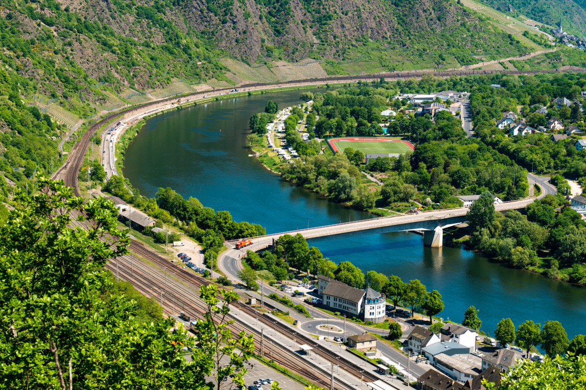 The Moselle River curves through Cochem, Germany, beneath steep vineyard-covered hills. A road bridge, railway tracks and a riverside sports ground are visible from the elevated viewpoint.