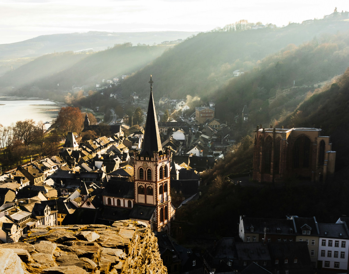 An elevated view overlooks the historic rooftops of Bacharach, Germany, with St Peter&rsquo;s Church and the Gothic Werner Chapel rising above the town. The Rhine and wooded valley hills extend into the distance.