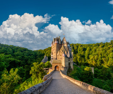 A stone bridge leads towards Eltz Castle near Wierschem, Germany. The medieval towers rise above the surrounding Eifel forest beneath a cloud-filled sky.