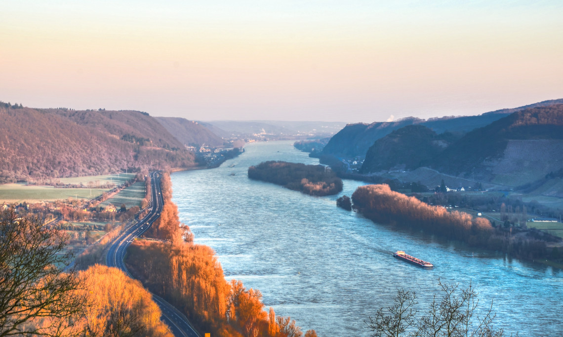 Scenic view over the Rhine River winding between wooded hills and riverside villages, with a cruise vessel sailing through the Upper Middle Rhine Valley.